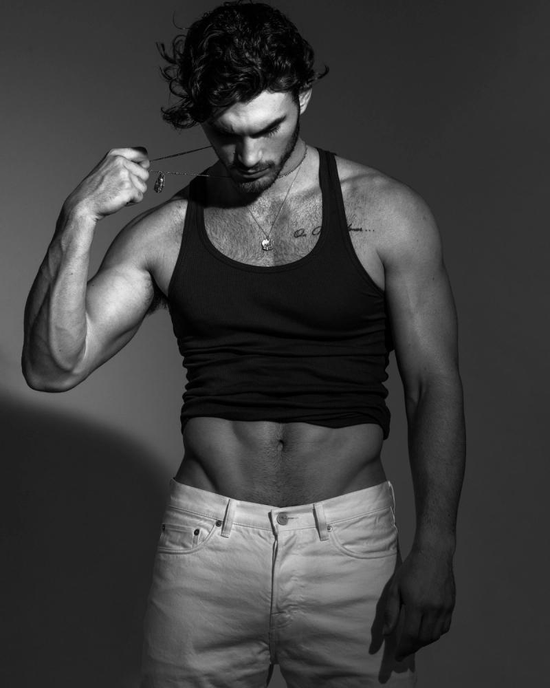 Michael Yerger in a black ribbed tank top and white jeans, tugging a silver necklace chain, for L'Officiel Hommes by photographer Taylor Miller.