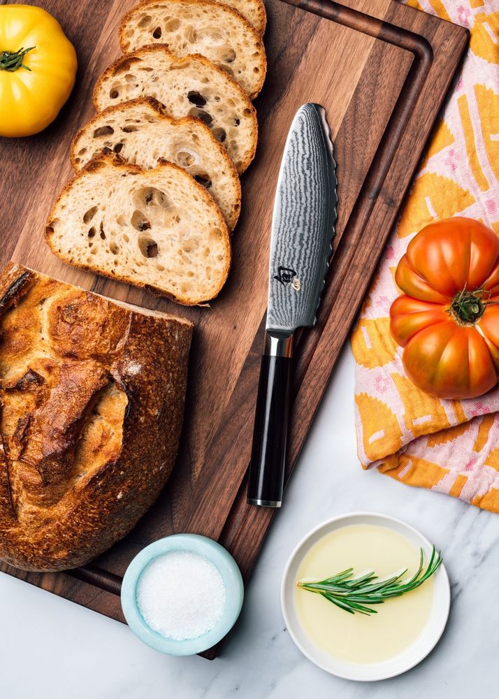 Kai damascus paring knife on a walnut board with sliced bread, a sourdough loaf, heirloom tomatoes, and rosemary oil, photographed for BuzzFeed by Taylor Miller.