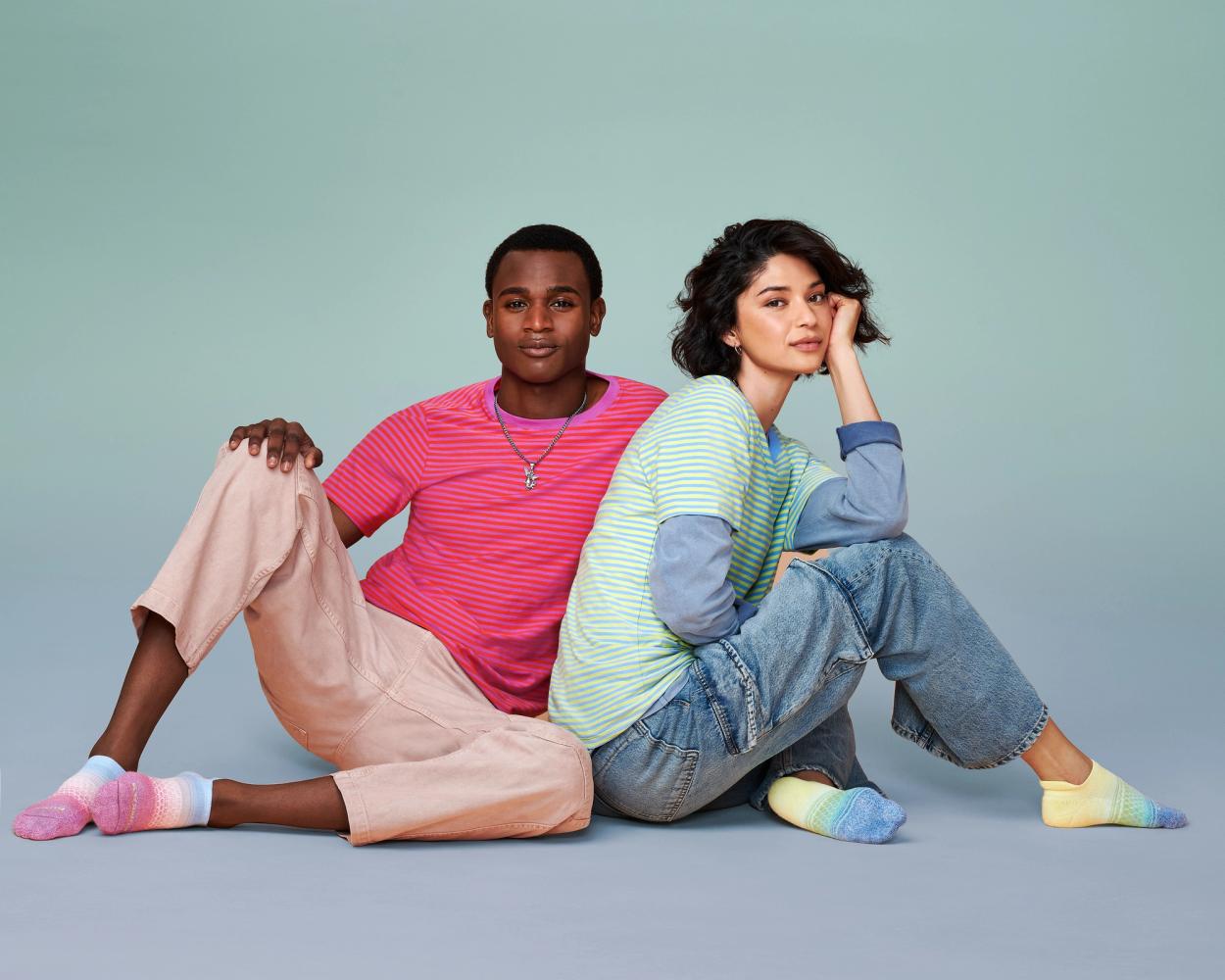 Four Dorzelma and Yared Chavez sit back-to-back in striped tees and denim, wearing rainbow gradient Bombas ankle socks, photographed for Bombas Pride 2024 by Taylor Miller.