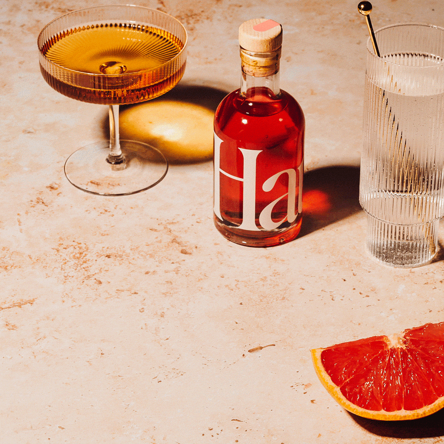 Sunlit cocktail flat lay with a red aperitif bottle, a coupe glass, a ribbed highball and grapefruit slices on warm stone, photographed by Taylor Miller.
