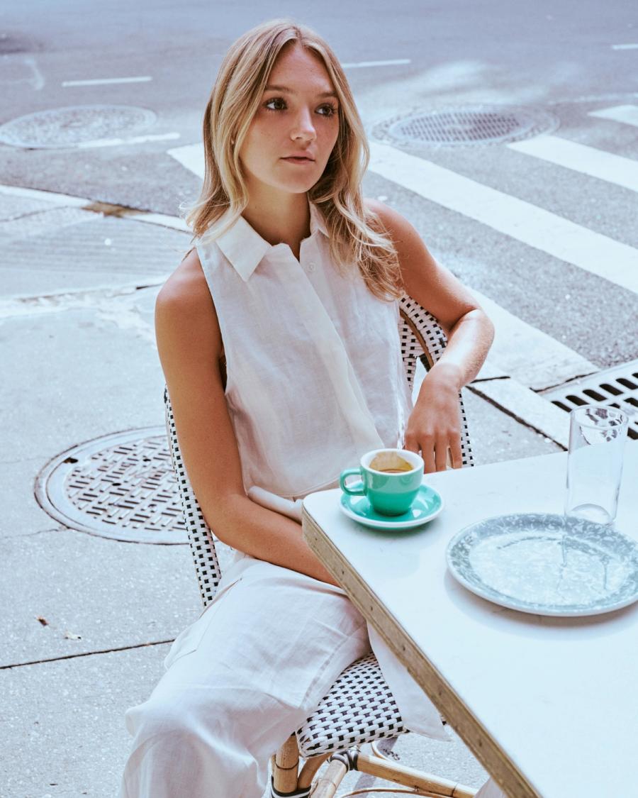 Julia Lisboa in a white sleeveless linen top seated at a sidewalk cafe table with an espresso, ESYA editorial campaign photographed by Taylor Miller.