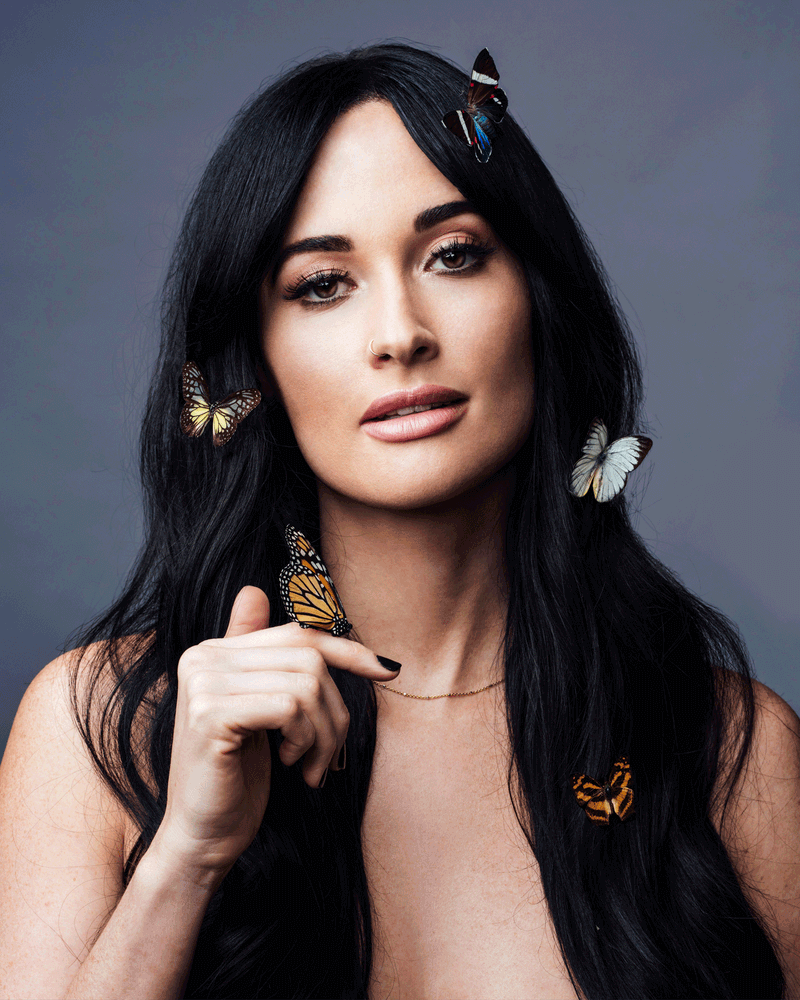 Kacey Musgraves with long dark hair as butterflies rest in her hair and on her hand, against a gray backdrop, photographed by Taylor Miller.