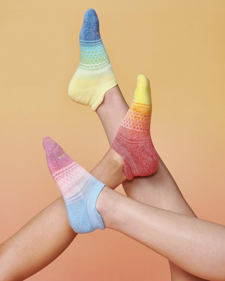 Three pairs of raised feet in rainbow gradient Bombas ankle socks against a peach backdrop, photographed for the Bombas Pride 2024 campaign by Taylor Miller.