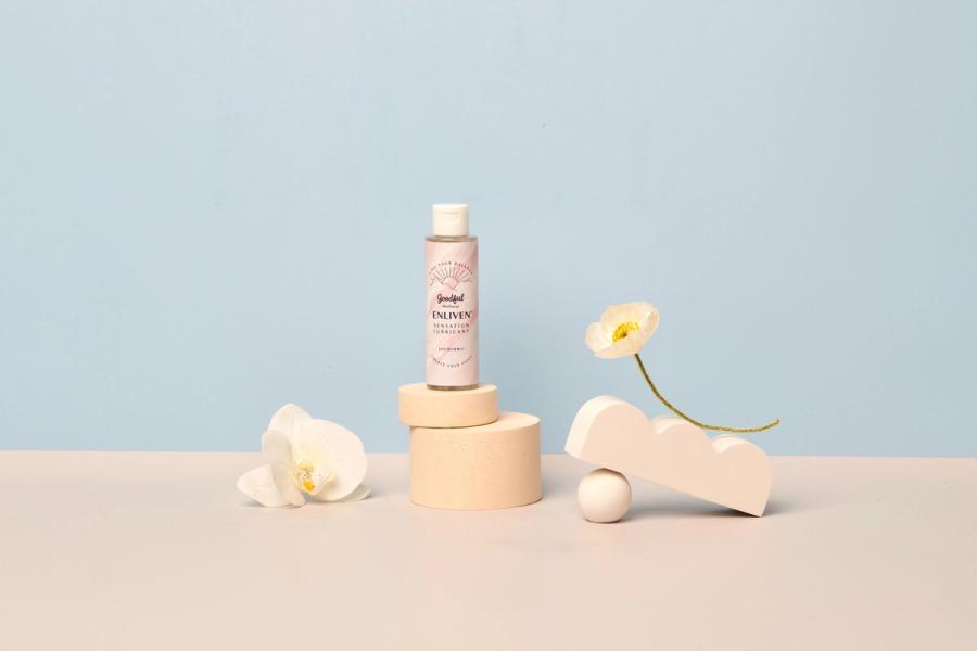 Goodful Wellness Enliven Sensation Lubricant on a pink plinth with orchid, poppy and white props against a blue backdrop, photographed for BuzzFeed by Taylor Miller.