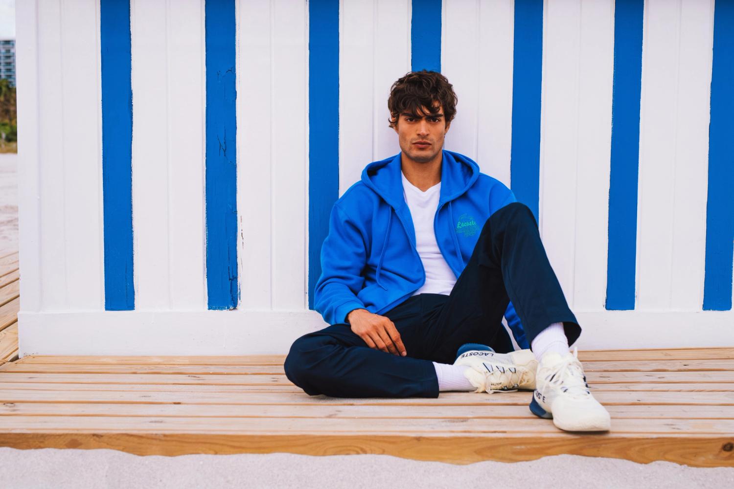 Iago Botelho in a blue Lacoste hoodie sits on a boardwalk by a striped beach wall, for Lacoste Miami Open 2025 by Taylor Miller.