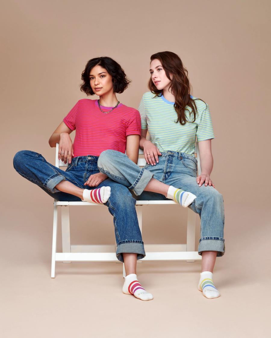 Yared Chavez and Francie Cohen in striped tees and cuffed jeans sit together wearing rainbow-striped Bombas ankle socks, photographed for the Bombas Pride 2024 campaign by Taylor Miller.