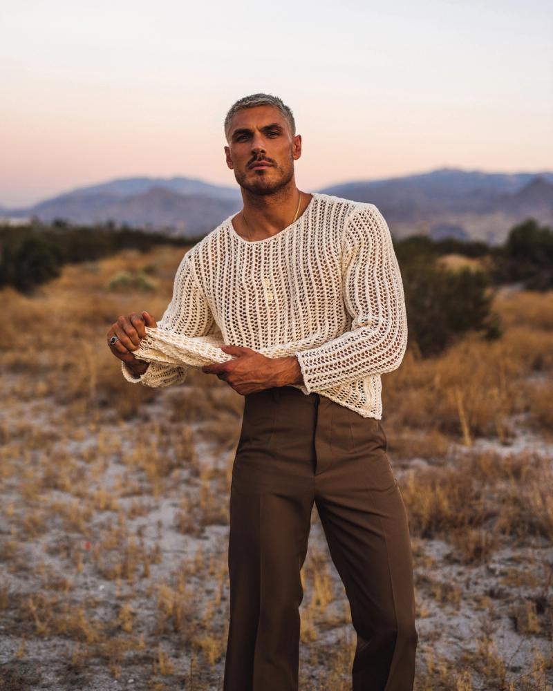 Chris Appleton in a cream open-knit sweater and brown trousers standing in a desert landscape at dusk, photographed by Taylor Miller.