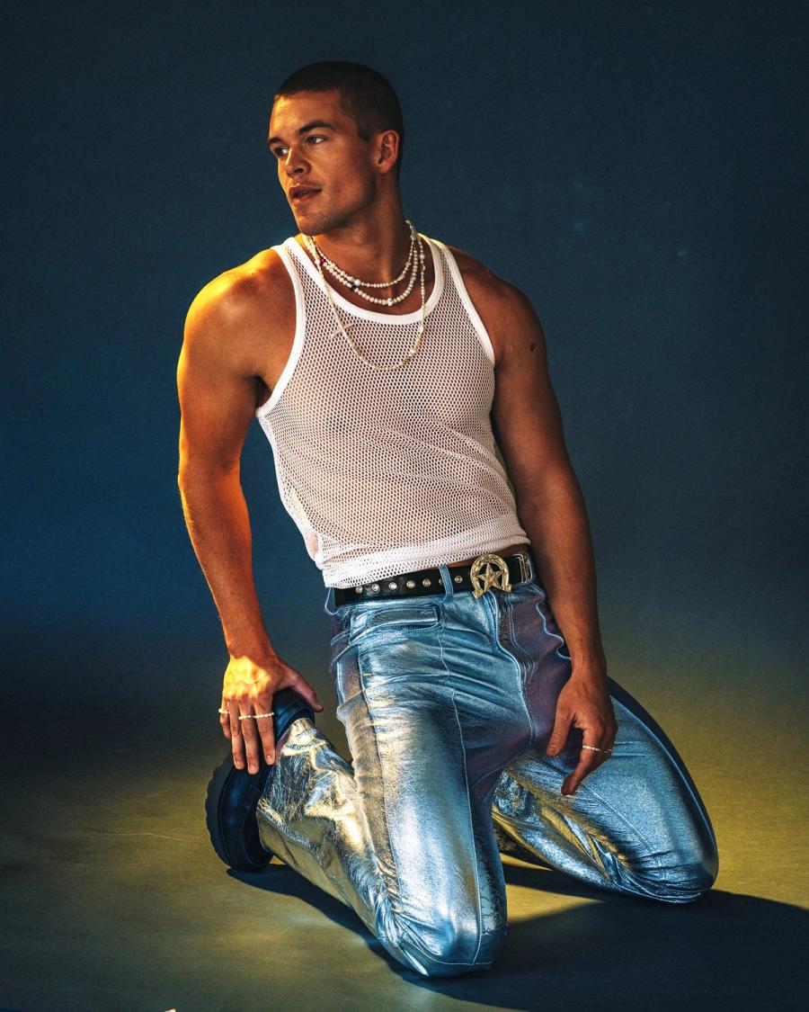Kneeling in a white mesh tank and metallic silver trousers with layered pearl necklaces, Mitchell Slaggert photographed under moody lighting for Numéro Netherlands by Taylor Miller.
