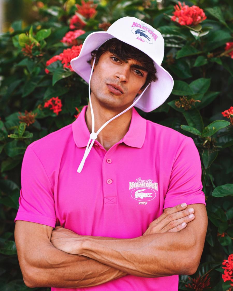 Iago Botelho in a hot pink Lacoste Miami Open 2025 polo and bucket hat poses before red blooms, photographed by Taylor Miller.