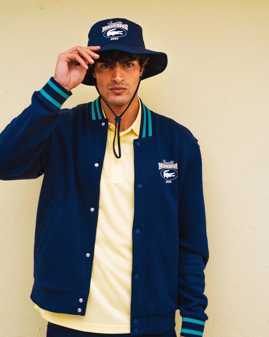 Iago Botelho tips a navy Lacoste Miami Open 2025 bucket hat over a varsity jacket and yellow polo, photographed by Taylor Miller.