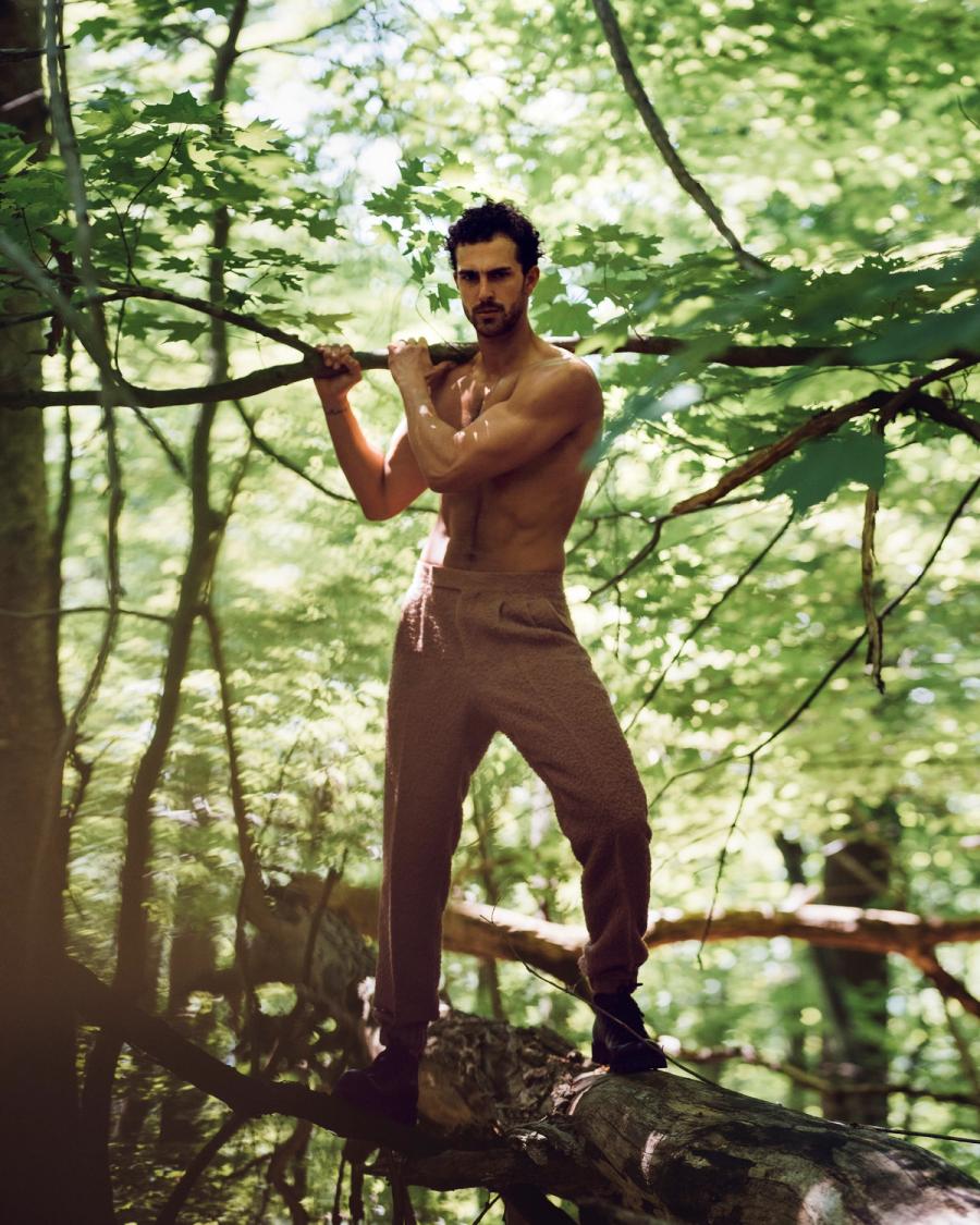 Clint Mauro in beige trousers gripping a branch while standing on a fallen tree in a green forest, photographed by Taylor Miller.