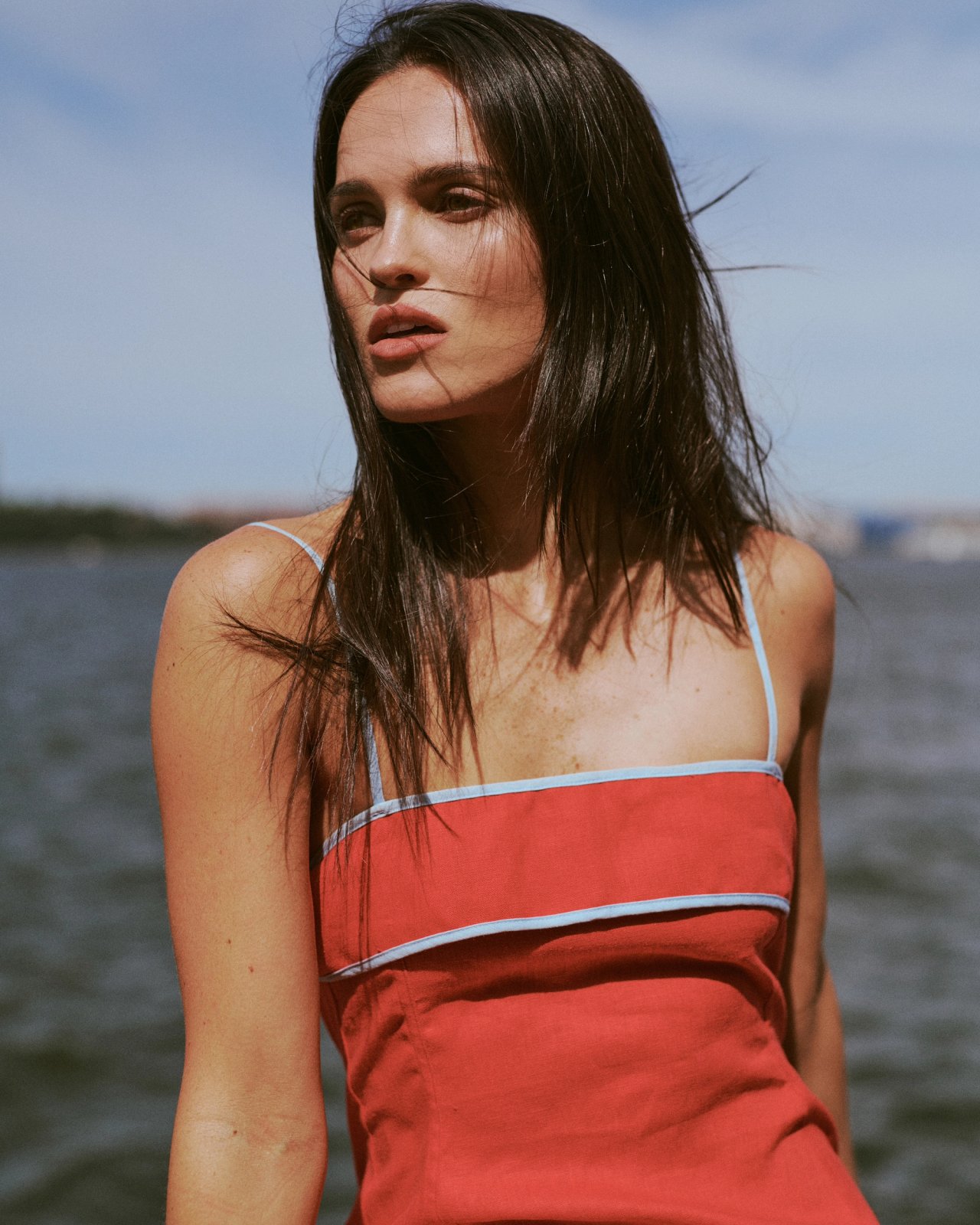 Mary Holland Nader in a red linen dress with blue trim poses by the water, ESYA editorial campaign photographed by Taylor Miller.