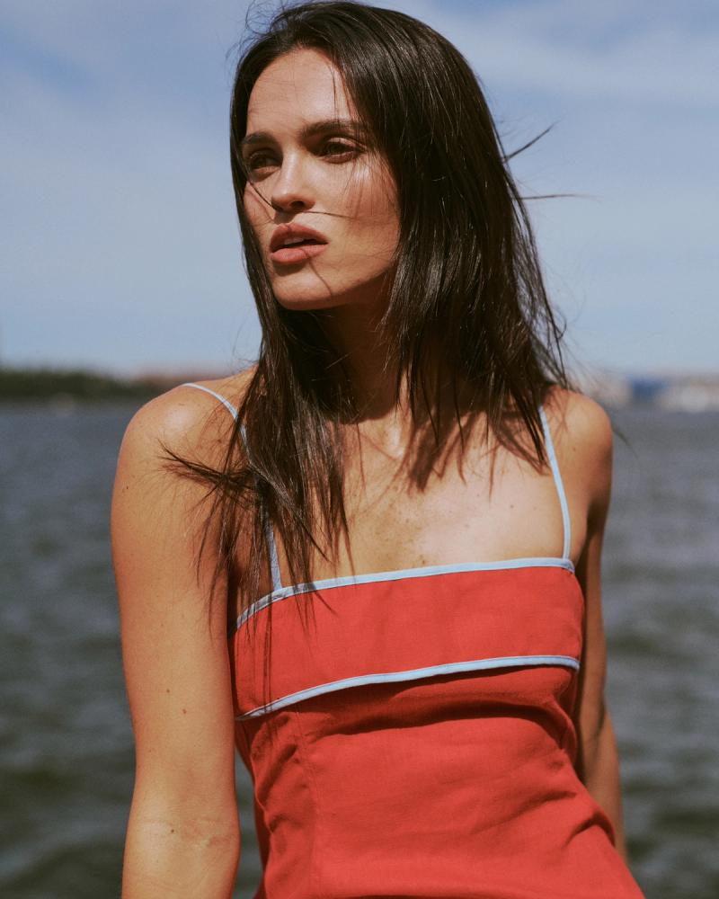 Mary Holland Nader in a red linen dress with blue trim poses by the water, ESYA editorial campaign photographed by Taylor Miller.