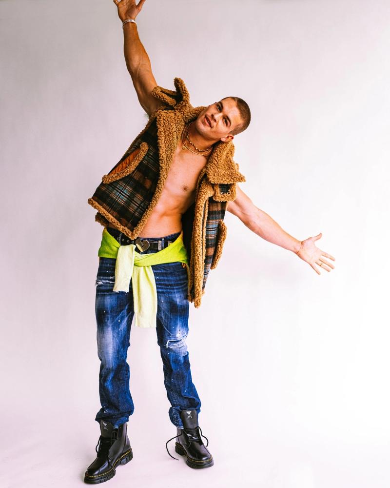 Arms outstretched, Mitchell Slaggert in a shearling-and-plaid jacket over a bare chest with distressed jeans and black boots, for Numéro Netherlands by Taylor Miller.