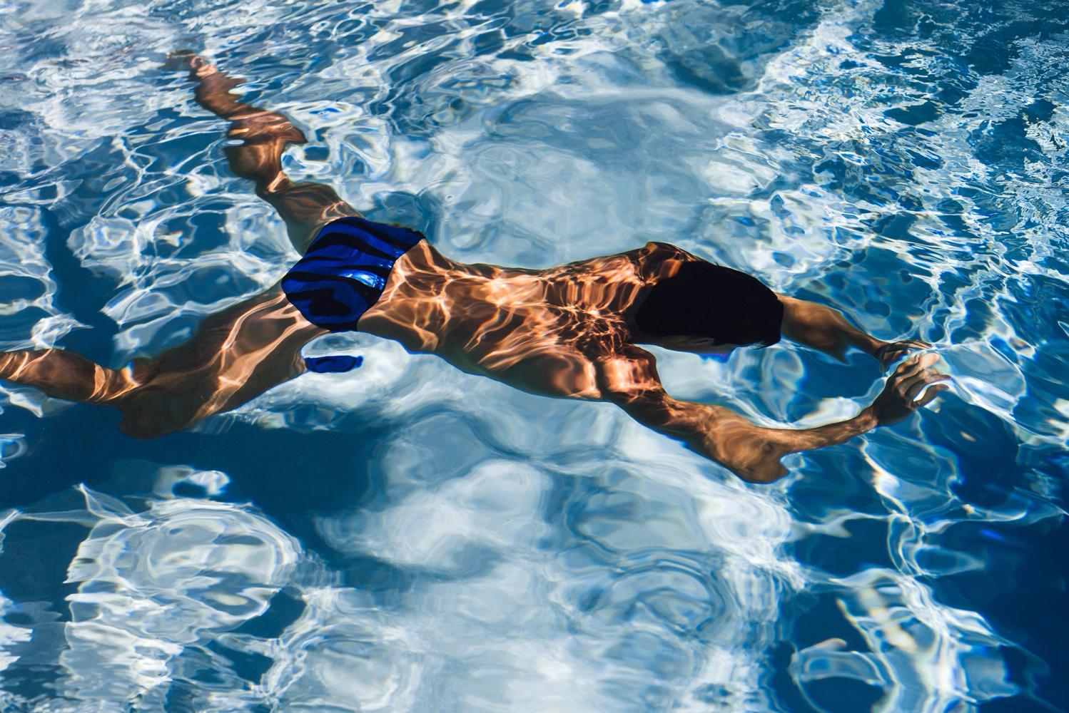 A male swimmer gliding underwater in blue trunks across a sunlit pool, photographed by Taylor Miller.