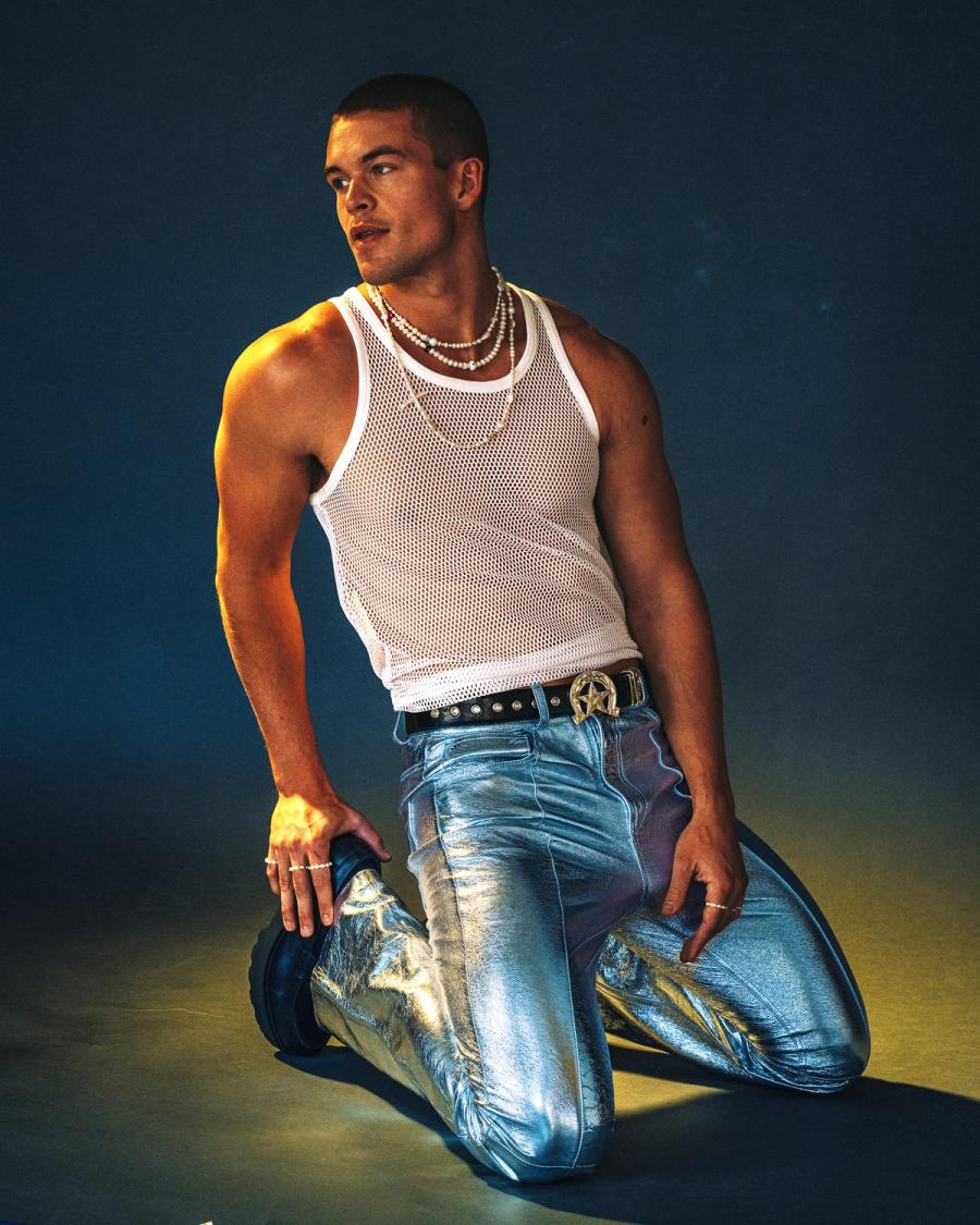 Mitchell Slaggert in a white mesh tank top and metallic silver trousers kneeling under moody studio light, photographed by Taylor Miller.