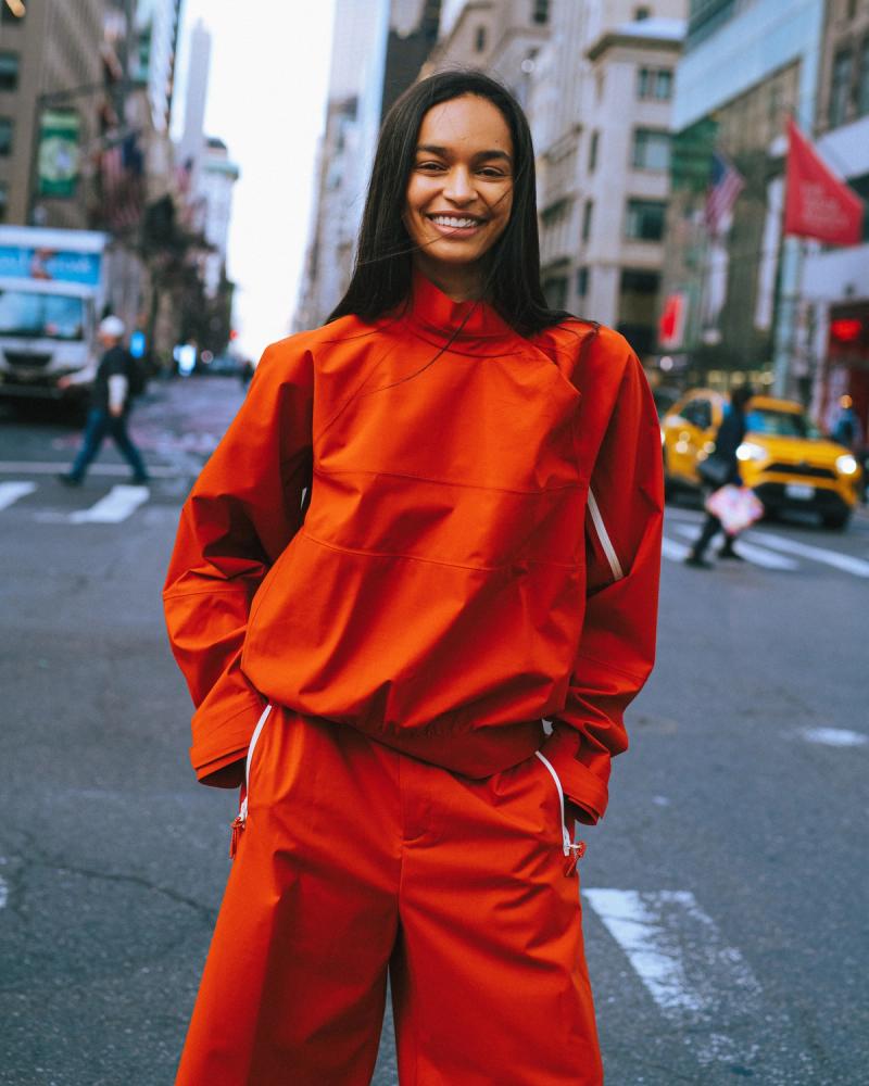 Franciele Santos in a bright orange Lacoste jacket and wide-leg trousers on a NYC street, for the Lacoste NYC 2026 campaign by Taylor Miller.