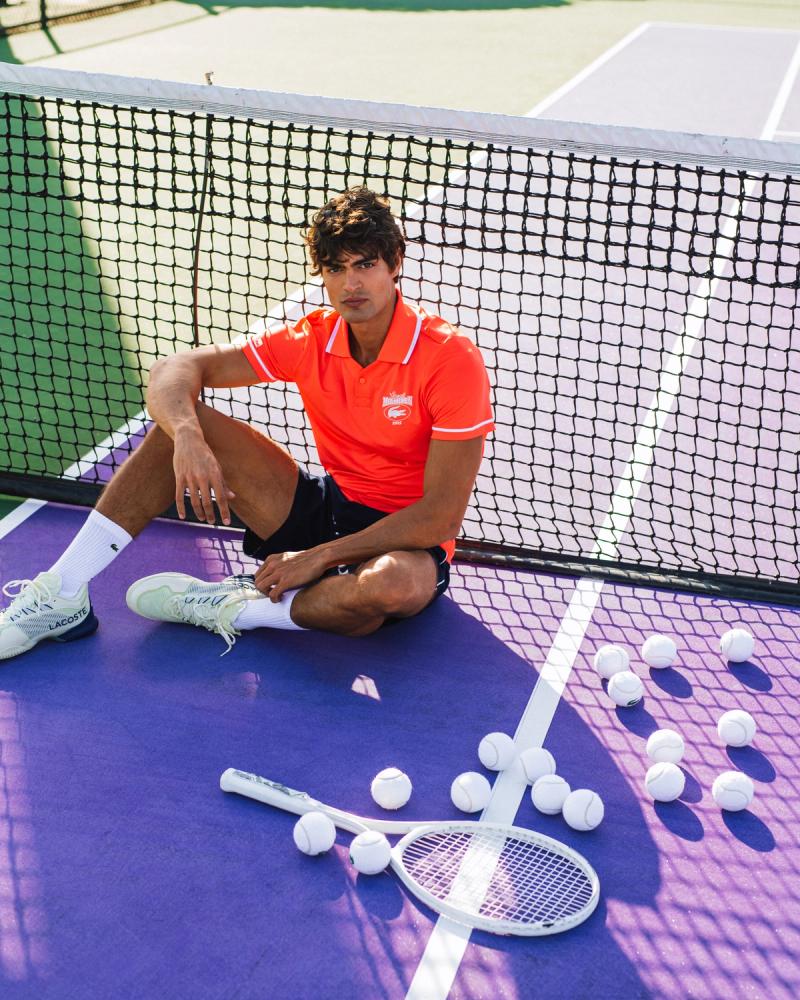 Iago Botelho in an orange Lacoste polo sits at the net amid tennis balls on a purple court, for Lacoste Miami Open 2025, photographed by Taylor Miller.