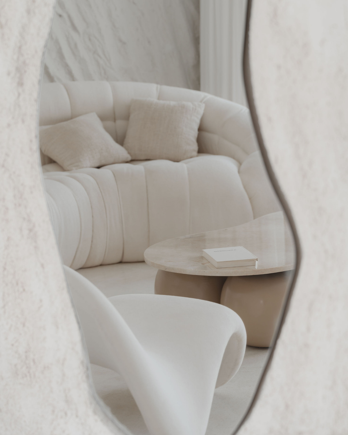 House of Creations Bali – Modern Lounge Interior View Through Mirror Reflection of curved white sofa and organic coffee table in designer lounge, showcasing minimal modern interior branding for House of Creations Bali villa