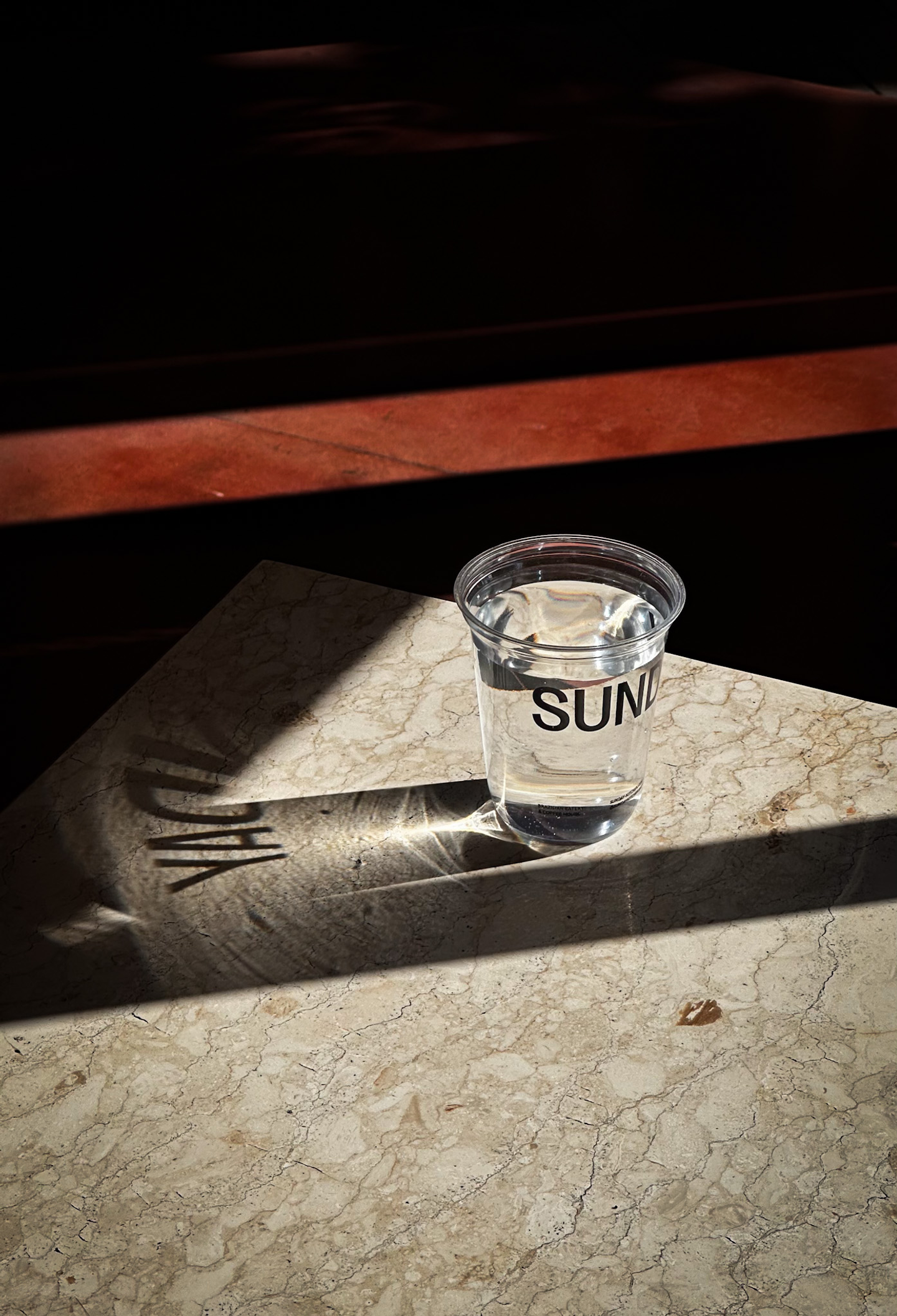 Stylish branded plastic water cup with “Sunday” logo on marble table in natural light, part of packaging design for Sunday Kitchen, Brazilian restaurant Canggu Bali
