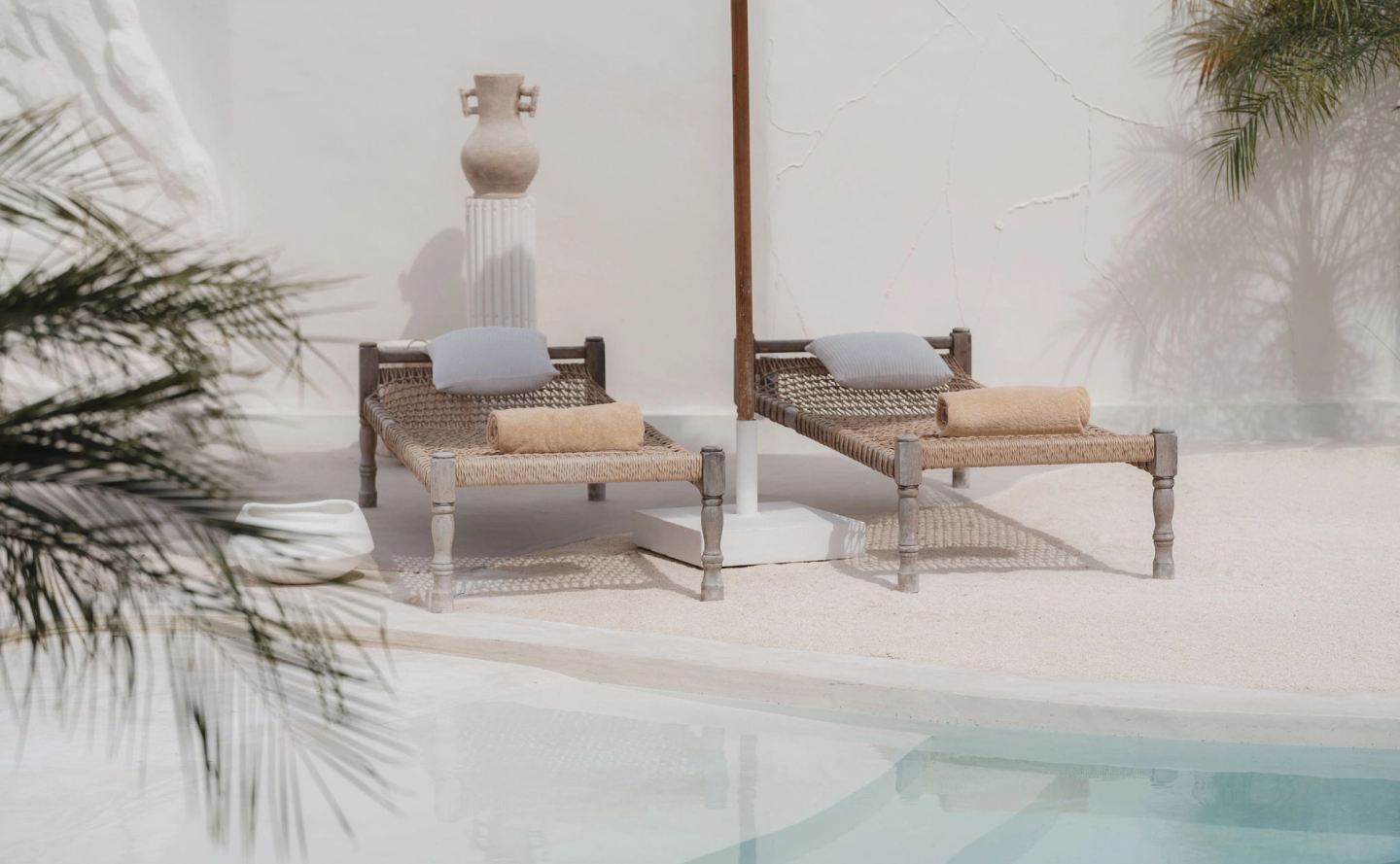 Twin woven sunbeds with pillows and towels beside pool, accented with sculptural ceramics and palm shadows, highlighting designer villa branding and leisure