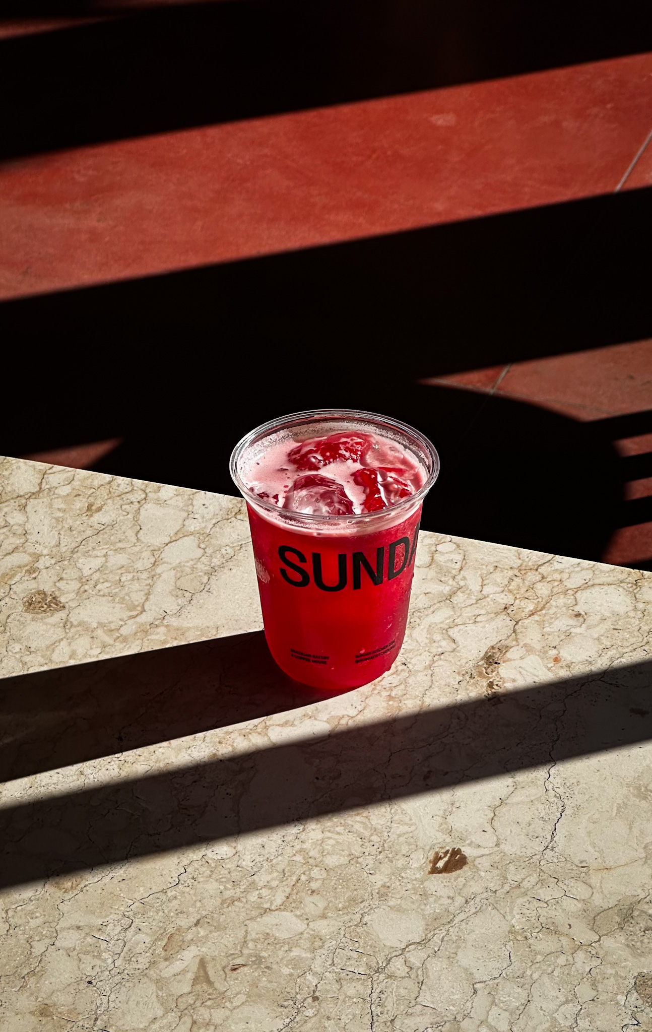 Sunday Kitchen Branded Beverage Cup – Packaging for Brazilian Restaurant in Bali Branded plastic beverage cup with “Sunday” logo and red drink on marble table, illustrating packaging design for Sunday Kitchen, Brazilian restaurant in Canggu Bali