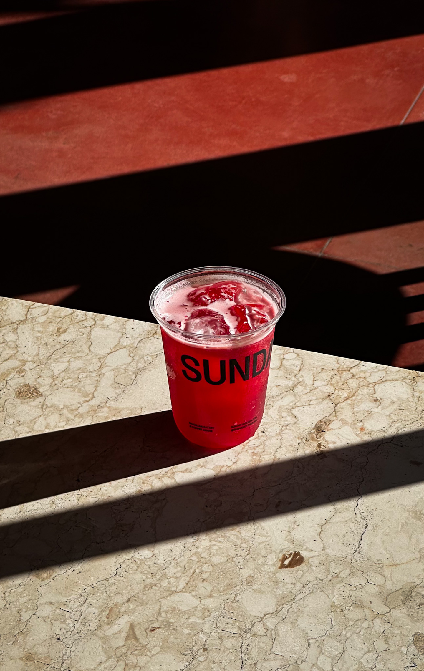 Sunday Kitchen Branded Beverage Cup – Packaging for Brazilian Restaurant in Bali Branded plastic beverage cup with “Sunday” logo and red drink on marble table, illustrating packaging design for Sunday Kitchen, Brazilian restaurant in Canggu Bali