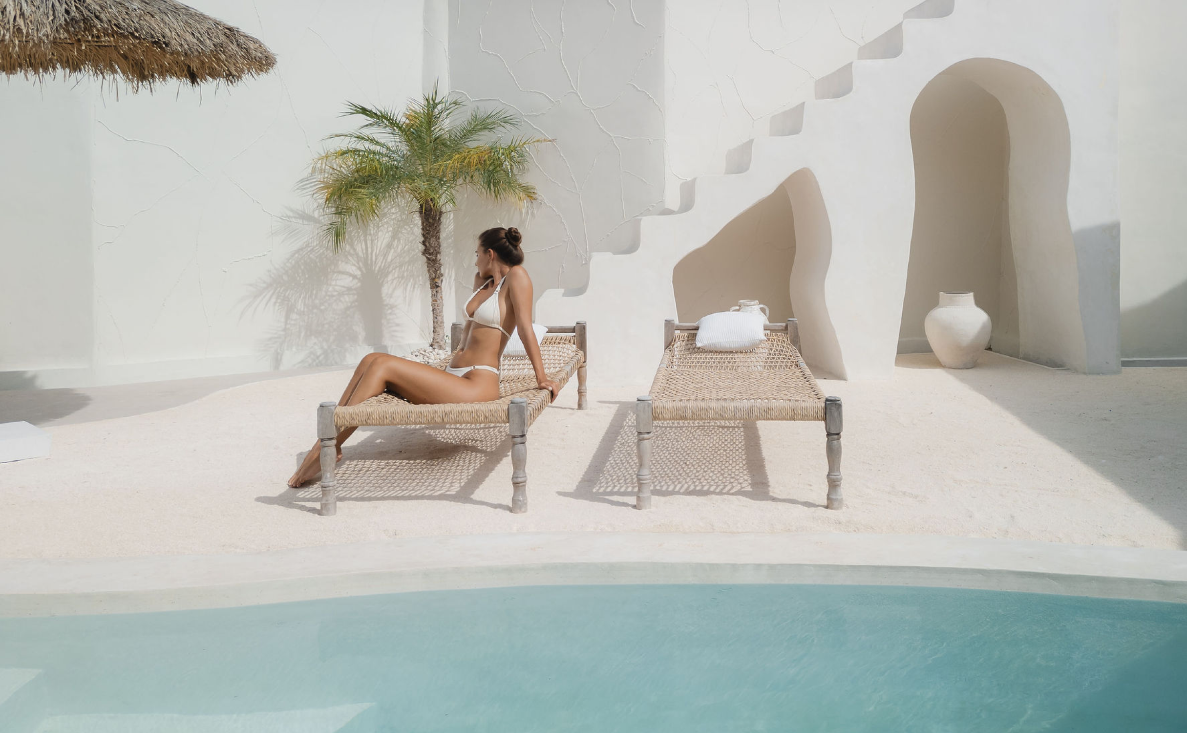 House of Creations Bali – Luxury Villa Poolside Relaxation Scene Woman in minimalist swimwear lounging on sunbed by pool with white modern villa background and palm, encapsulating Bali designer villa lifestyle for House of Creations