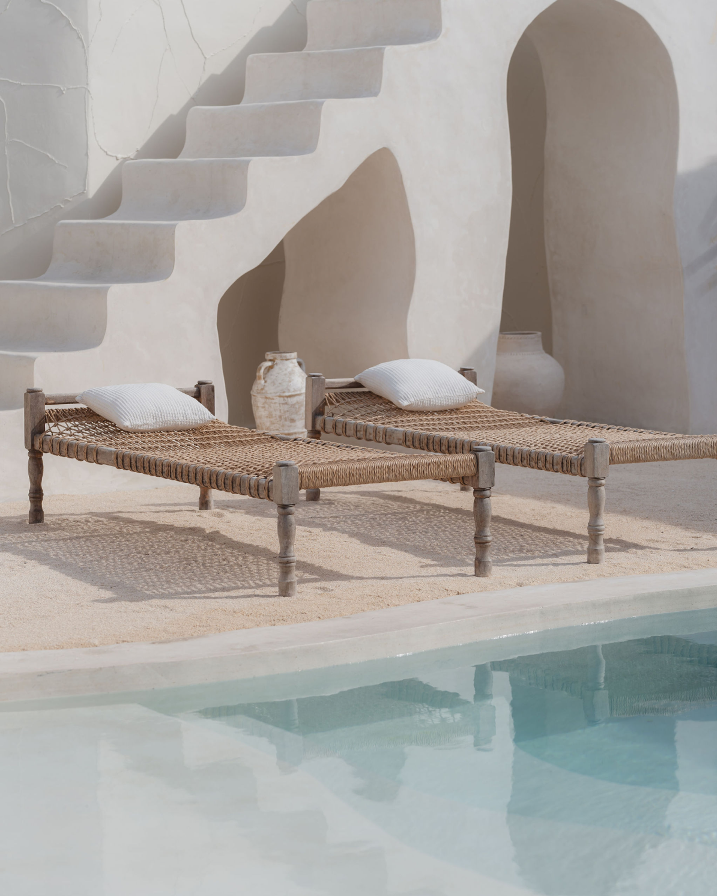 House of Creations Bali – Poolside Designer Sunbeds and Stairs Woven sunbeds with pillows beside pool, with sculptural stone stairs and arched wall, luxury boutique villa branding for House of Creations Bali