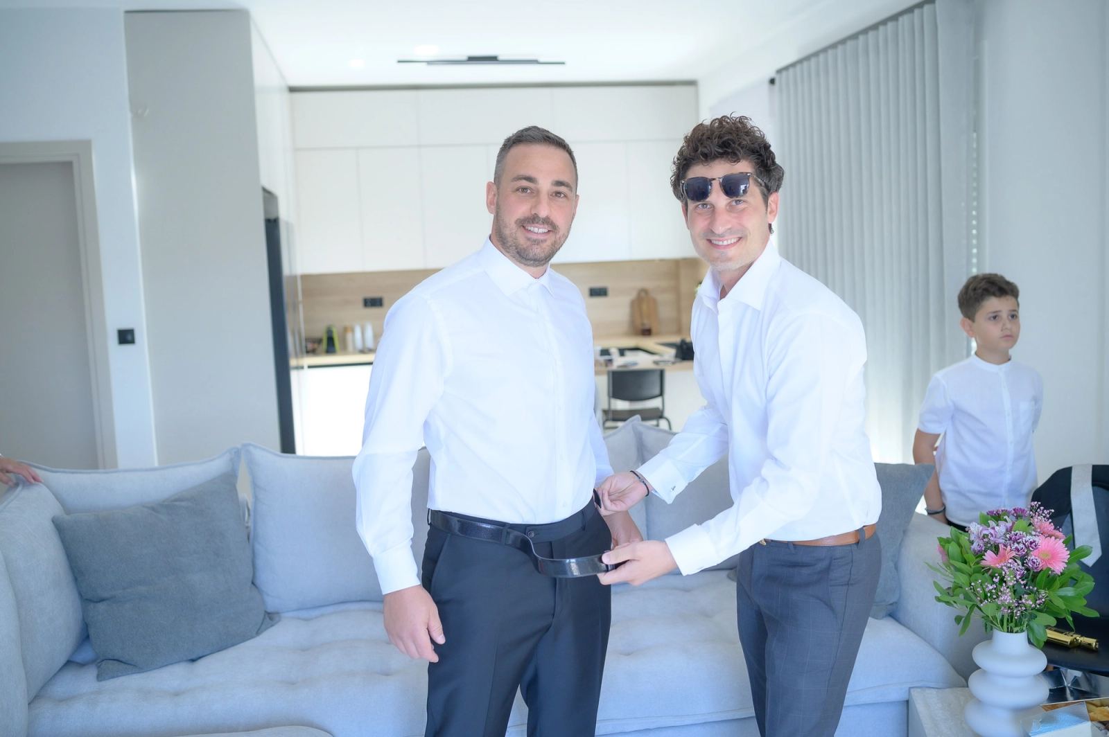 A man in sunglasses helps the groom with his belt in a bright living room.