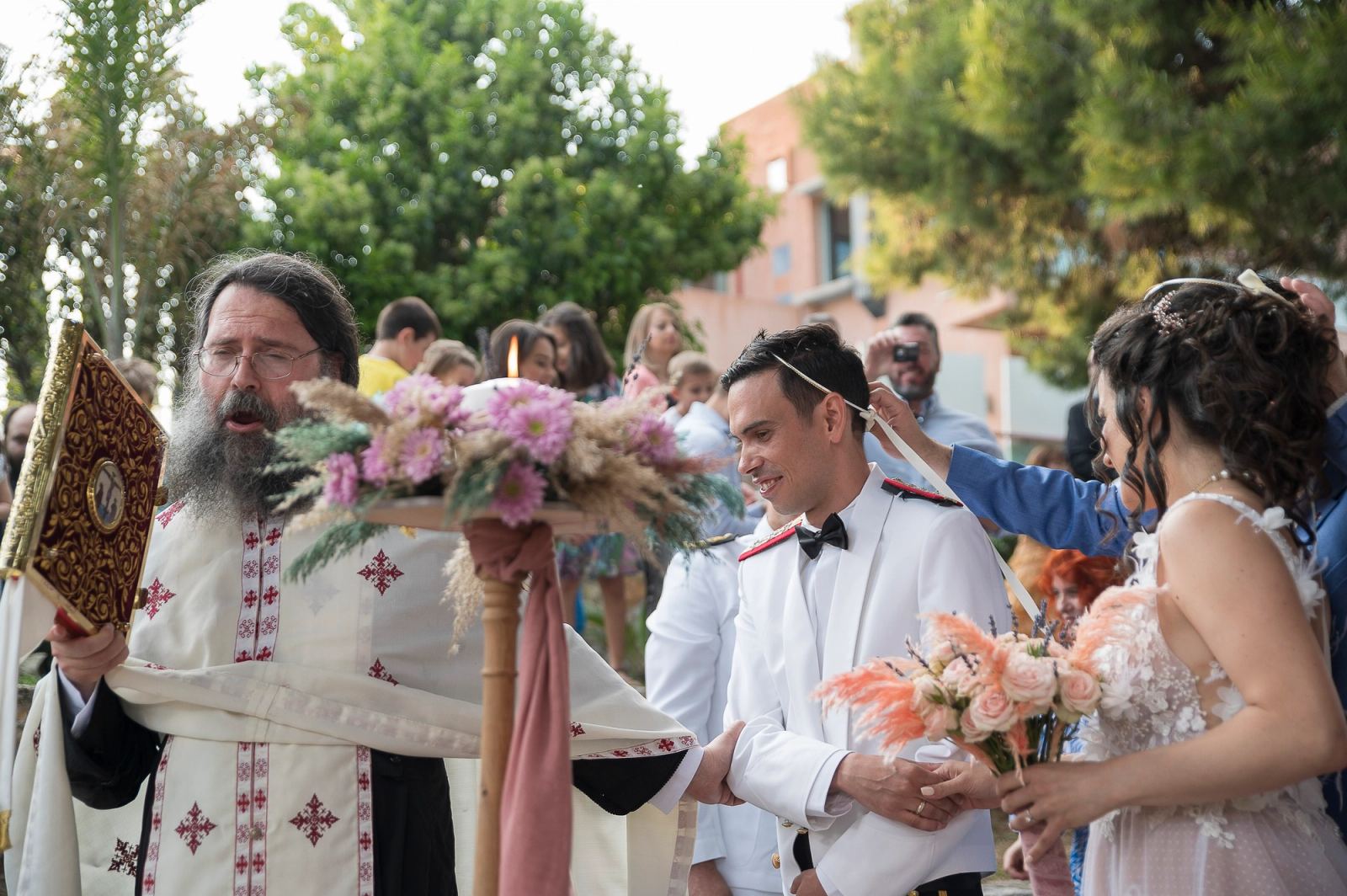 Ο ιερέας, ο γαμπρός με λευκή στολή και η νύφη περπατούν στον χορό του Ησαΐα, με καλεσμένους πίσω.