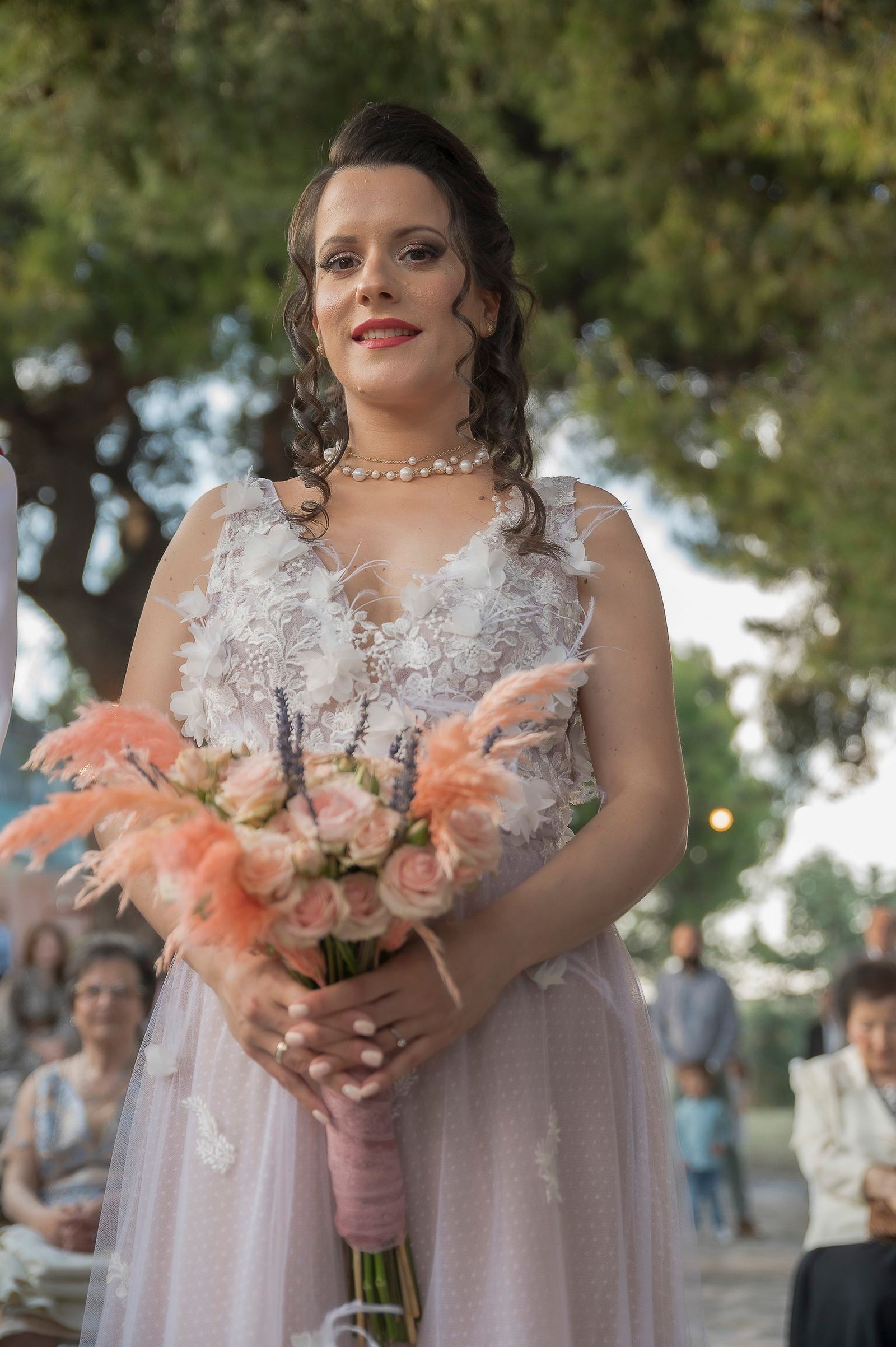 Η νύφη κρατά ροζ ανθοδέσμη μπροστά της, με δέντρα και καλεσμένους στο φόντο.