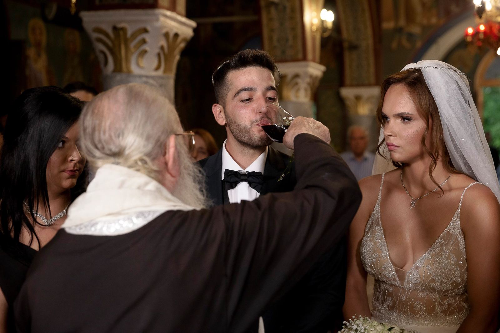 Ο ιερέας δίνει στον γαμπρό να πιει από το κοινό ποτήριο, ενώ η νύφη στέκεται δίπλα.