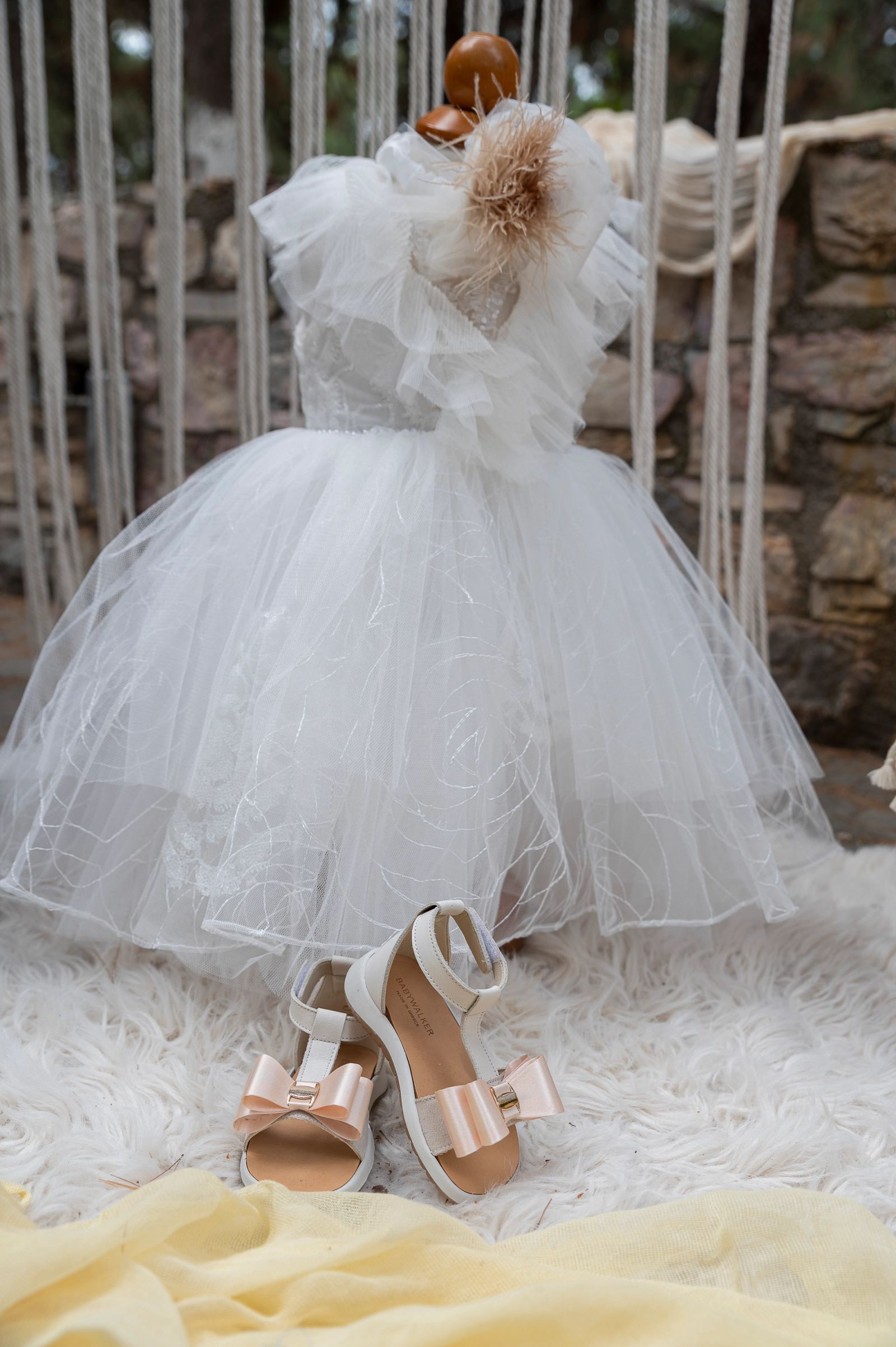 White tulle christening dress on a stand and white sandals with pink bows on a fluffy rug.