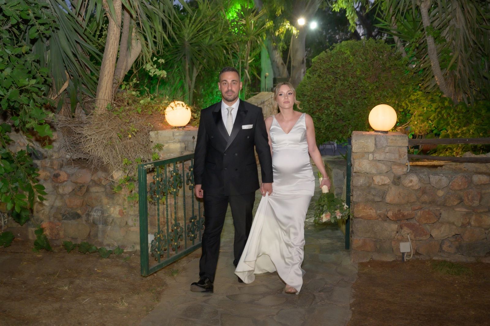 The bride and groom walk through an iron gate on a stone path lined with globe lights and green-lit plants.