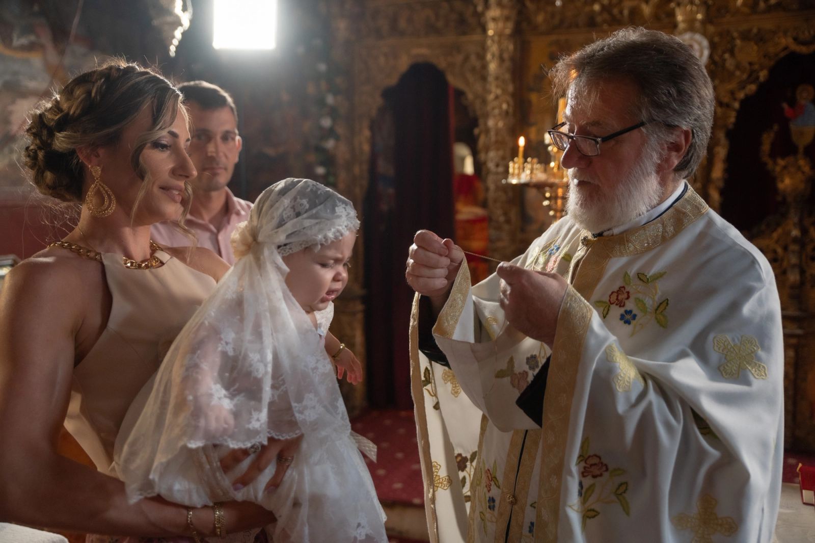 The priest holds a cord before a woman carrying the baby in a lace veil, a gilded iconostasis behind.