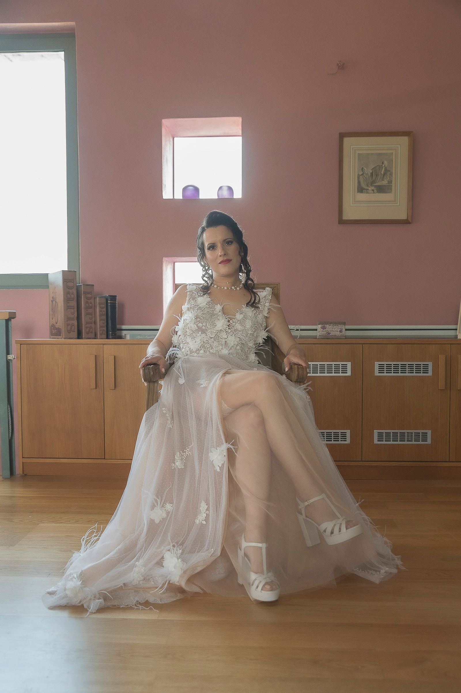 Η νύφη κάθεται σε πολυθρόνα με σταυρωμένα πόδια, με νυφικό από τούλι και λευκά πέδιλα, σε ροζ δωμάτιο.