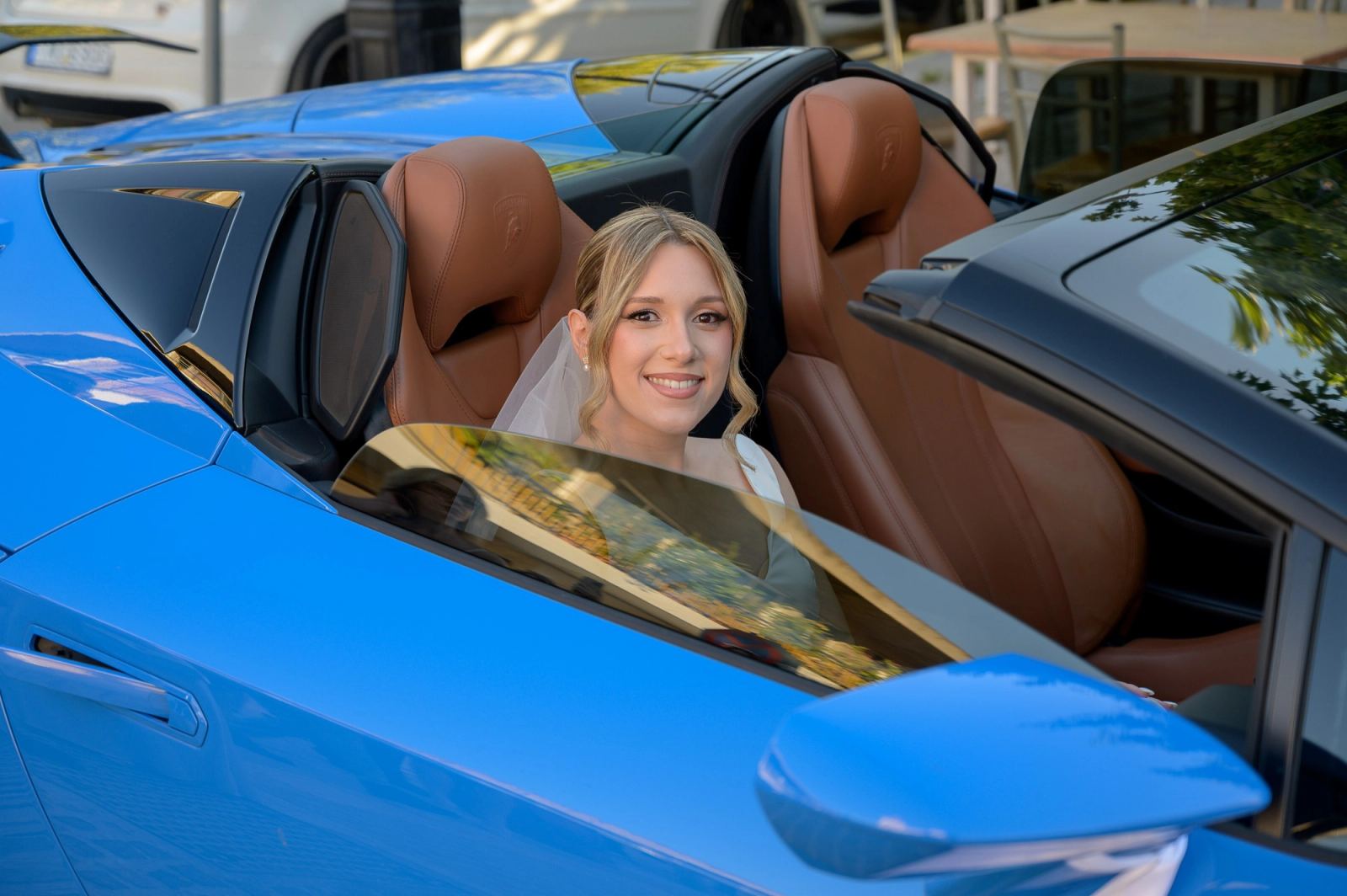 The bride in a veil sits smiling in a blue open-top sports car with tan leather seats.