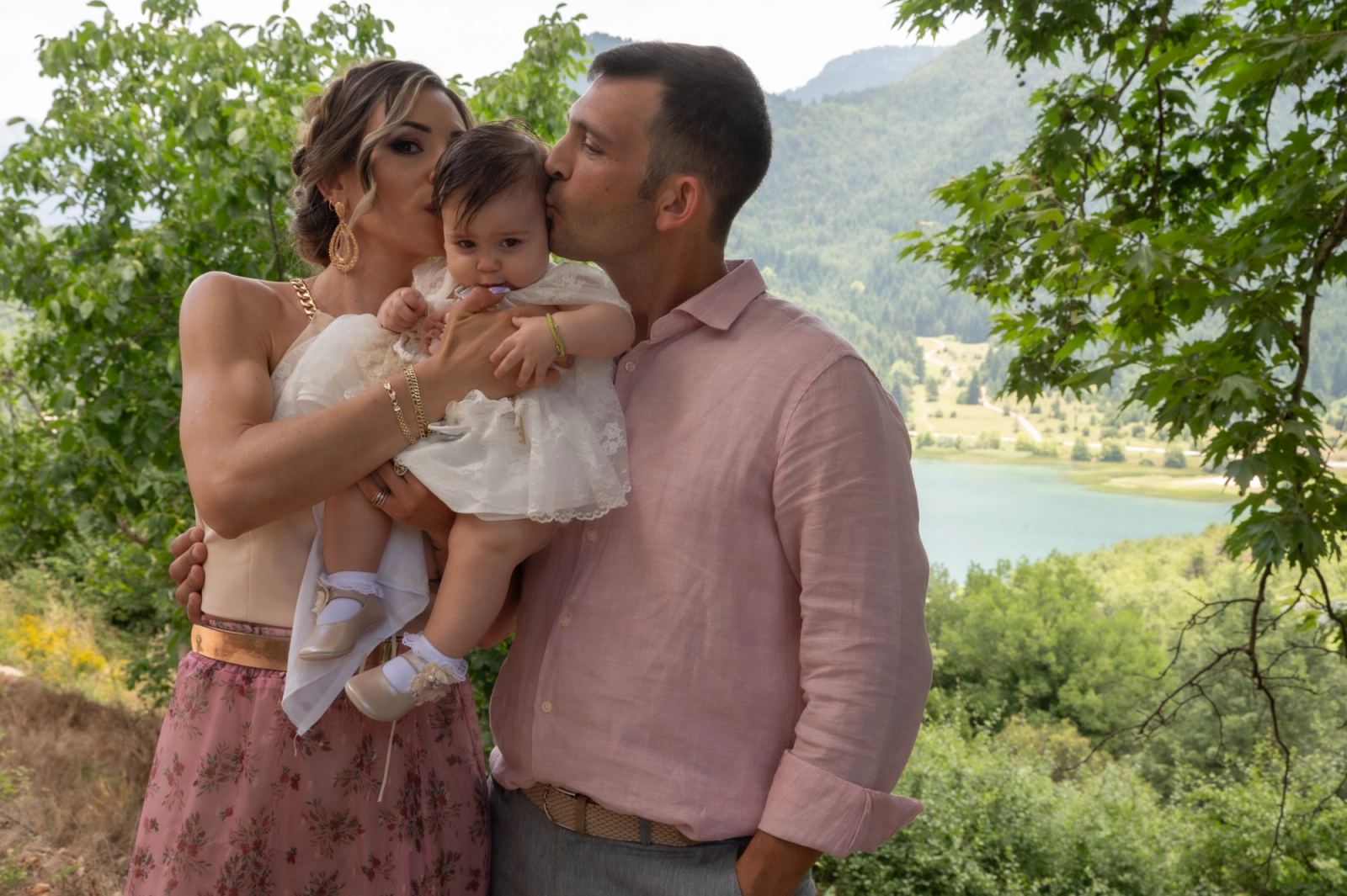 A man and a woman kiss the baby in white with a lake and green mountains behind.
