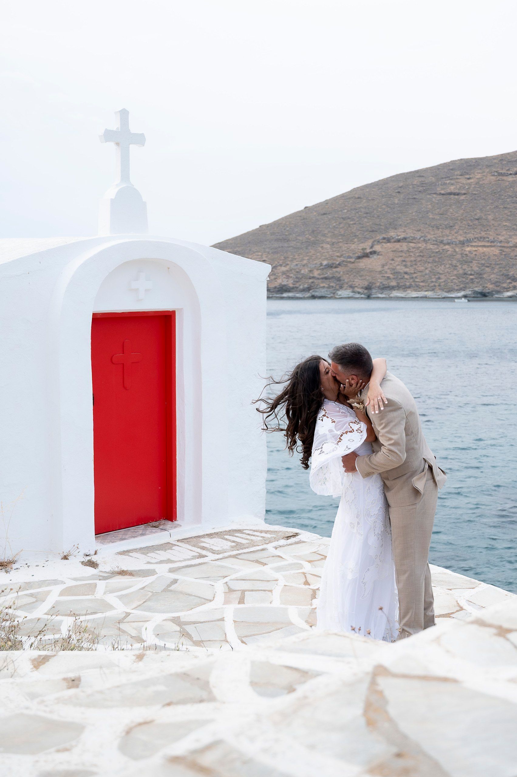 Η νύφη και ο γαμπρός φιλιούνται δίπλα σε άσπρο εκκλησάκι με κόκκινη πόρτα, με θάλασσα και λόφο πίσω τους.