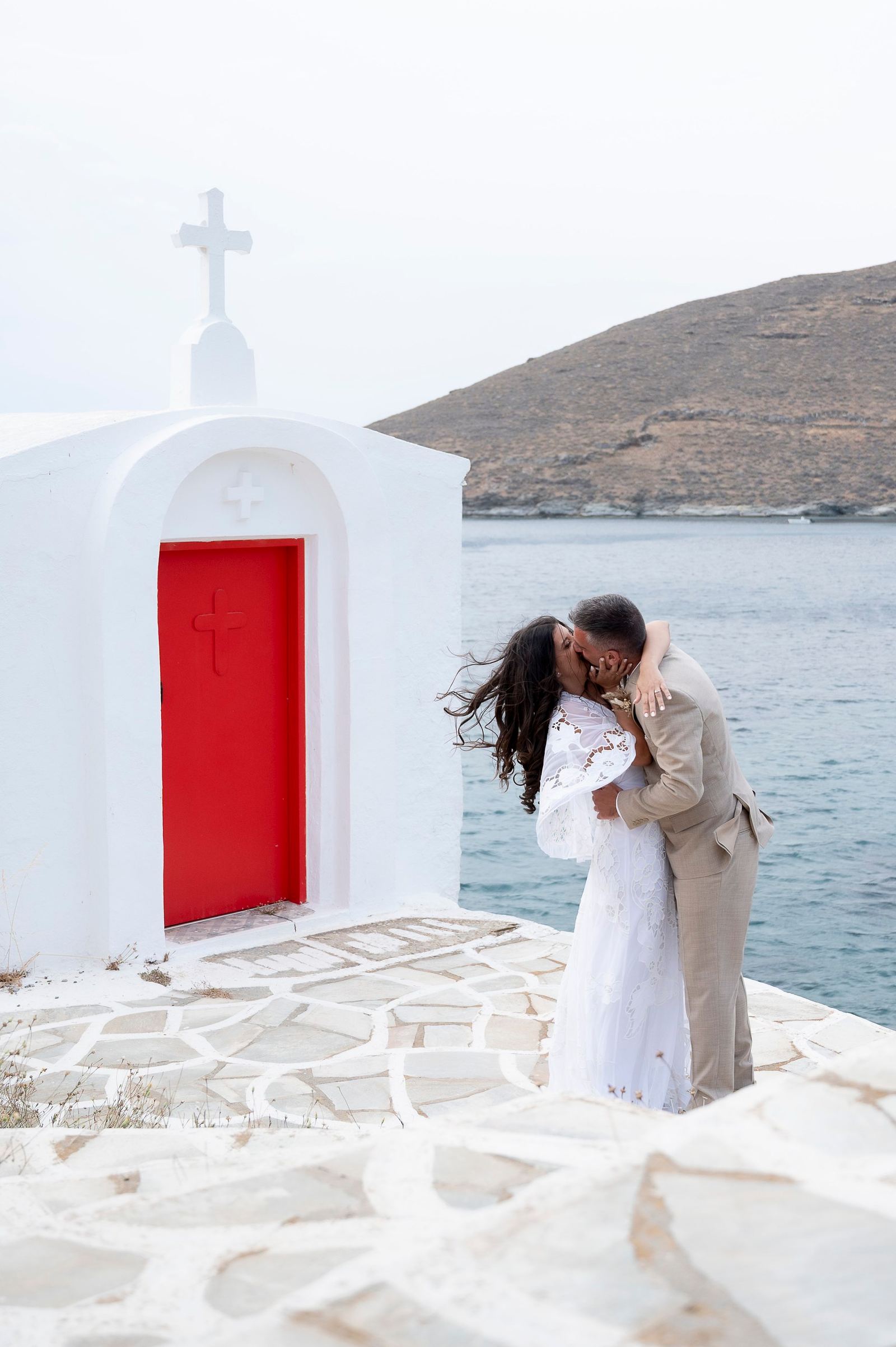 Η νύφη και ο γαμπρός φιλιούνται δίπλα σε άσπρο εκκλησάκι με κόκκινη πόρτα, με θάλασσα και λόφο πίσω τους.