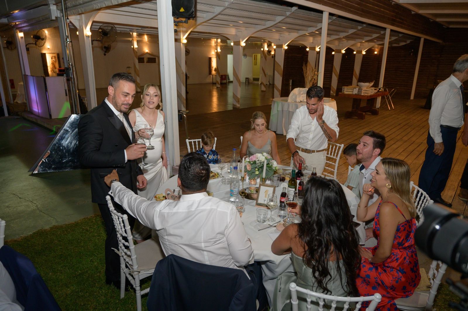 The groom and bride, holding glasses, stand beside a table of guests at the outdoor reception.