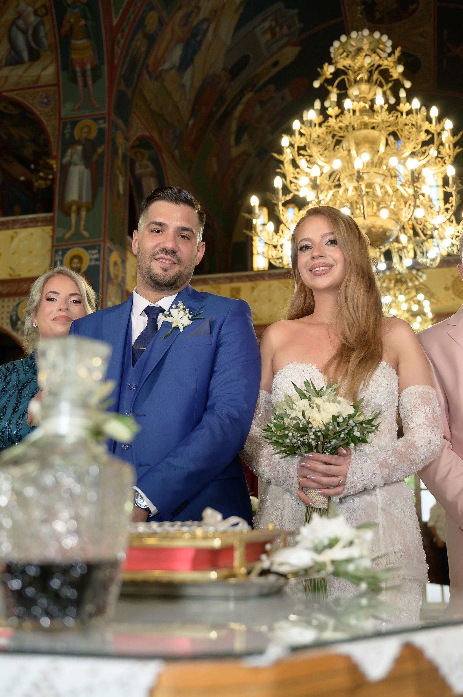 Ο γαμπρός με μπλε κοστούμι και η νύφη με ανθοδέσμη στέκονται κάτω από χρυσό πολυέλαιο σε εκκλησία με αγιογραφίες.