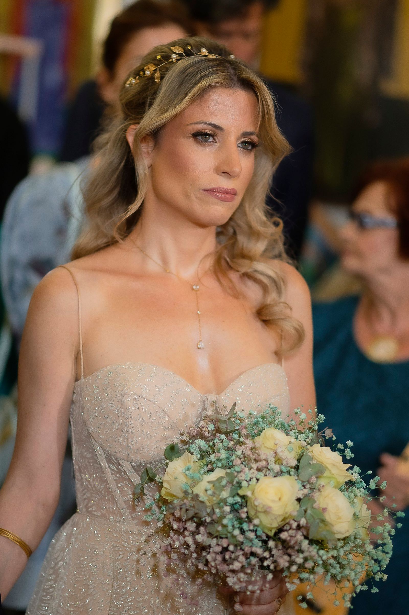 Η νύφη με χρυσή στέκα και ανθοδέσμη στέκεται ανάμεσα σε καλεσμένους στην εκκλησία.