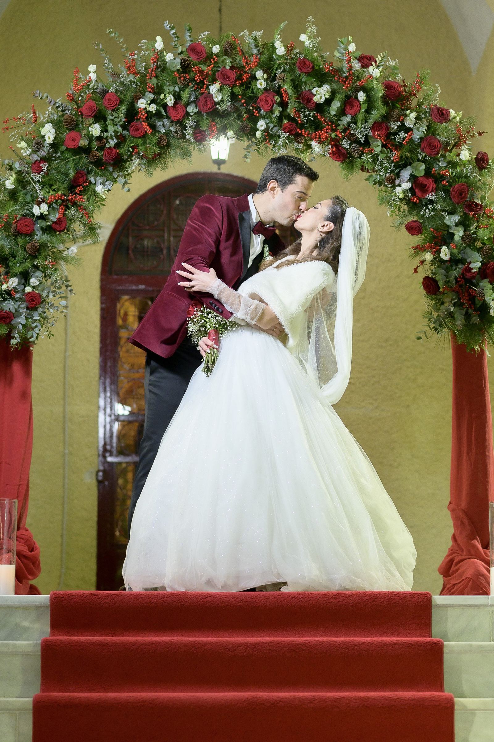 The bride and groom kiss under an arch of red roses above red-carpeted steps