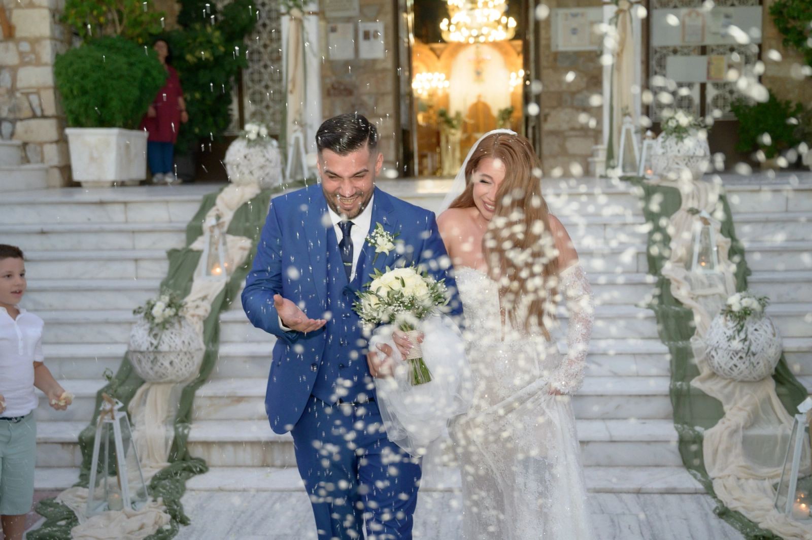 Ο γαμπρός και η νύφη στα σκαλιά της εκκλησίας μέσα σε βροχή από ρύζι, με ένα αγόρι στα αριστερά.