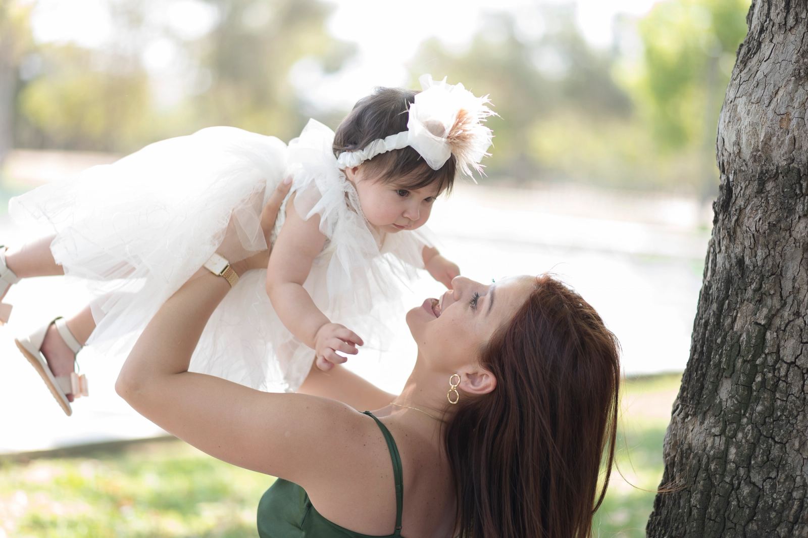 A woman in a green dress lifts the baby in a white dress high beside a tree trunk.