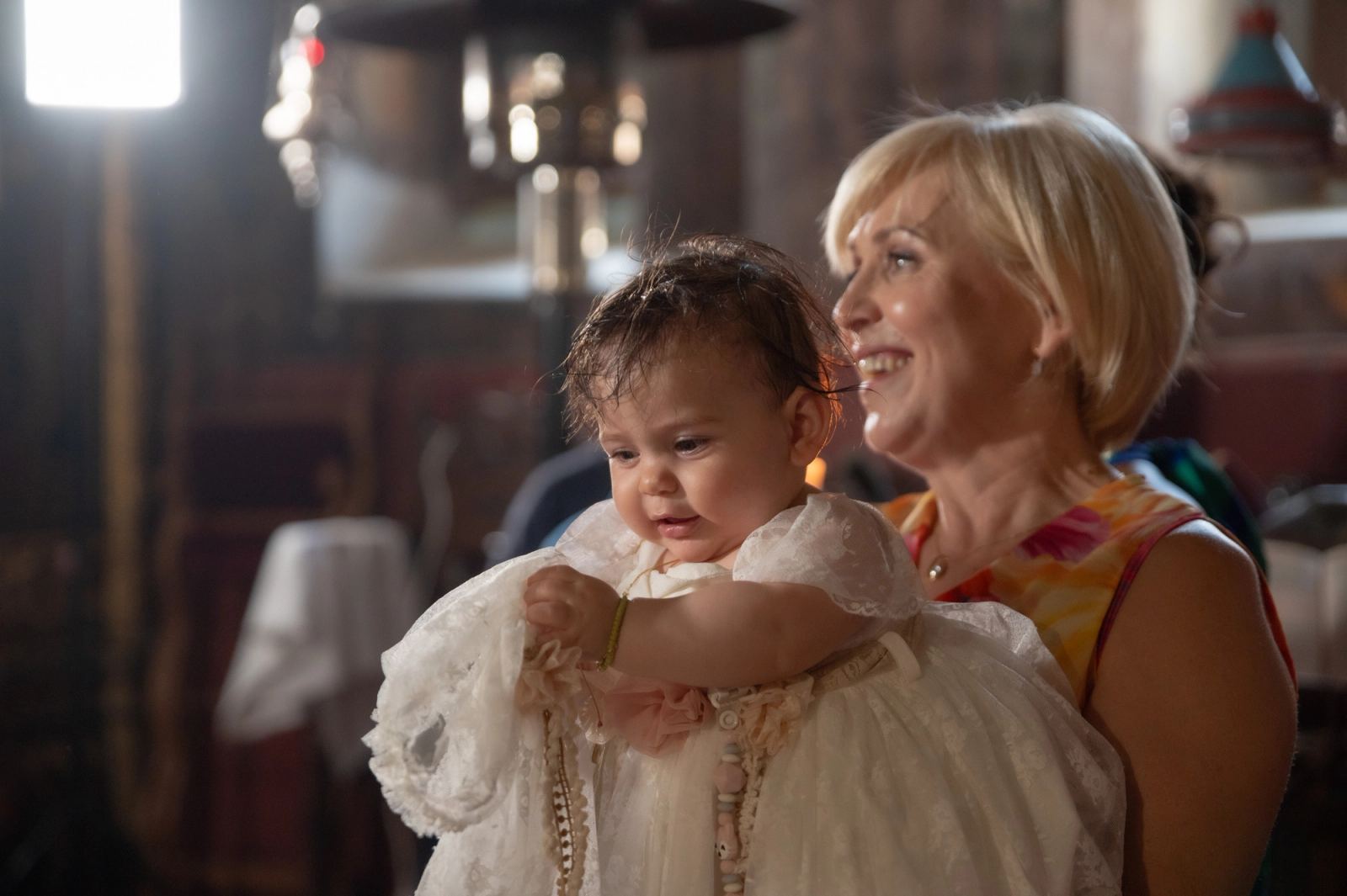 A blonde woman smiles holding the baby in a white lace dress inside the church.