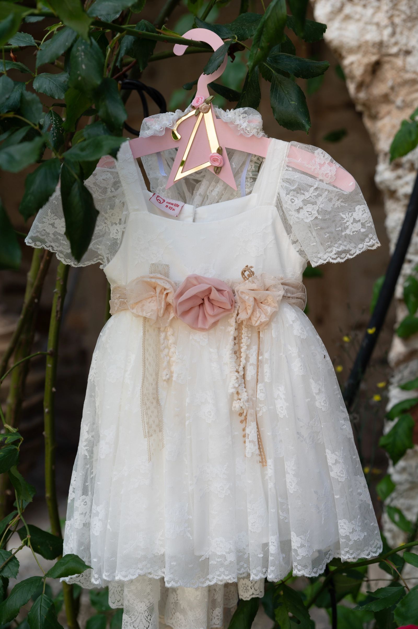 A white lace christening dress with pink flowers hangs on a pink hanger among leaves.