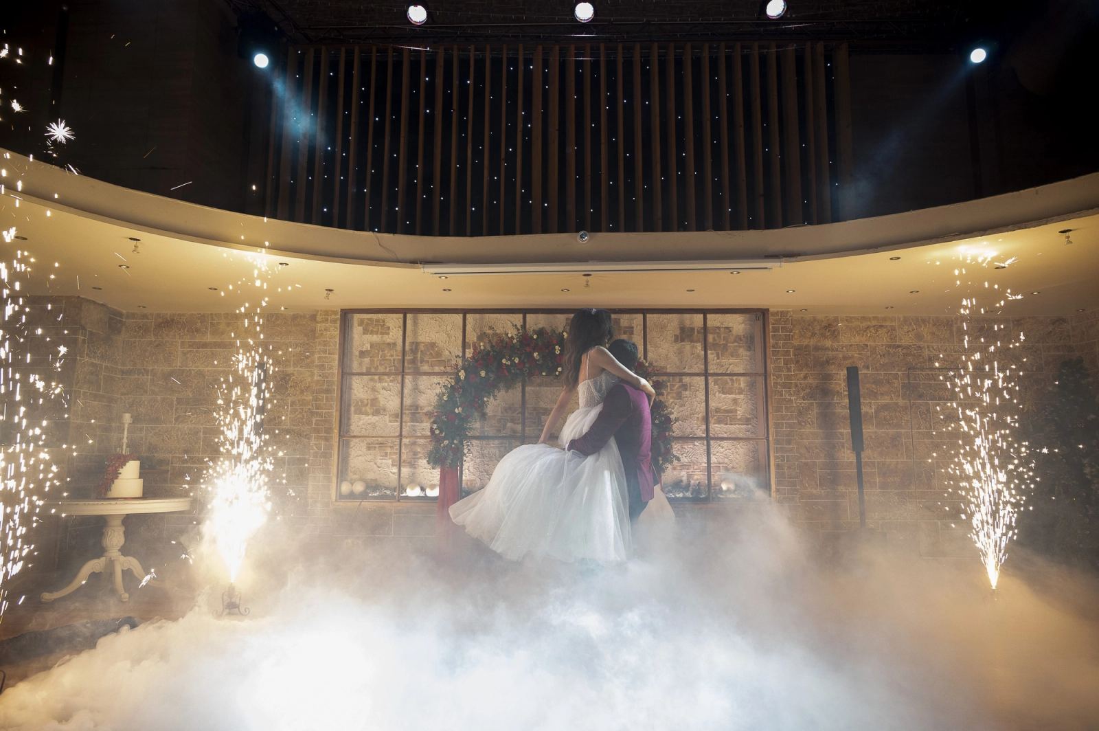 The groom lifts the bride during the dance between spark fountains and low fog in a reception hall.